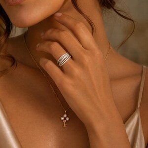 Pode incluir: Uma mão usando um anel de diamante de várias faixas. Um colar de ouro com um pingente de chave também é visível. As joias são colocadas em um fundo macio e neutro, destacando o brilho dos diamantes.