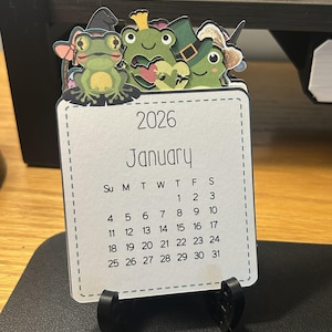 May include: A 2026 desk calendar featuring a January calendar page. The top of the calendar has a decorative element with three cartoon frogs wearing witch hats. The calendar is supported by a black stand.