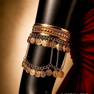 May include: A gold-toned arm cuff featuring a wide band with a pyramid stud texture. Two rows of dangling gold coin charms are connected by delicate chains, creating a layered effect that encircles the arm. The piece has a polished metallic finish.