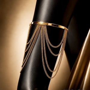 May include: A gold-toned arm cuff featuring a smooth, solid band that encircles the arm, adorned with multiple delicate, dangling gold chains that drape downwards in layered, cascading loops. The text INKORACLE ADORNMENT is visible at the bottom.