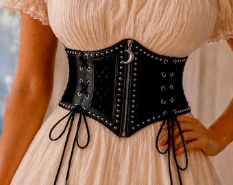 May include: A black corset belt featuring a front zipper closure with a silver crescent moon charm, silver stud detailing along the edges, and black lace-up side panels with long dangling cords. The corset is worn over a cream off-the-shoulder dress.