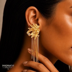 Puede incluir: Pendiente dorado con diseño floral, un cristal central y flecos de cadena colgantes. El pendiente se lleva en la oreja, mostrando sus intrincados detalles y su elegante estilo. La marca "INKORACLE - ADORNMENT" es visible en la parte inferior.