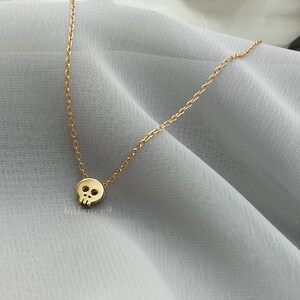 Puede incluir: Un delicado collar dorado con un peque&ntilde;o colgante de calavera. La calavera tiene cuencas de los ojos y una cavidad nasal. La cadena es fina y dorada, y el collar se muestra sobre una tela gris claro.