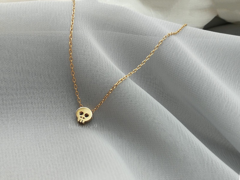 Puede incluir: Un delicado collar dorado con un peque&ntilde;o colgante de calavera. La calavera tiene cuencas de los ojos y una cavidad nasal. La cadena es fina y dorada, y el collar se muestra sobre una tela gris claro.