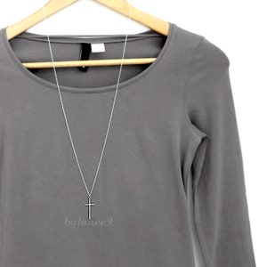 May include: A silver chain necklace with a simple silver cross pendant hanging from it. The necklace is displayed on a grey long-sleeved shirt.