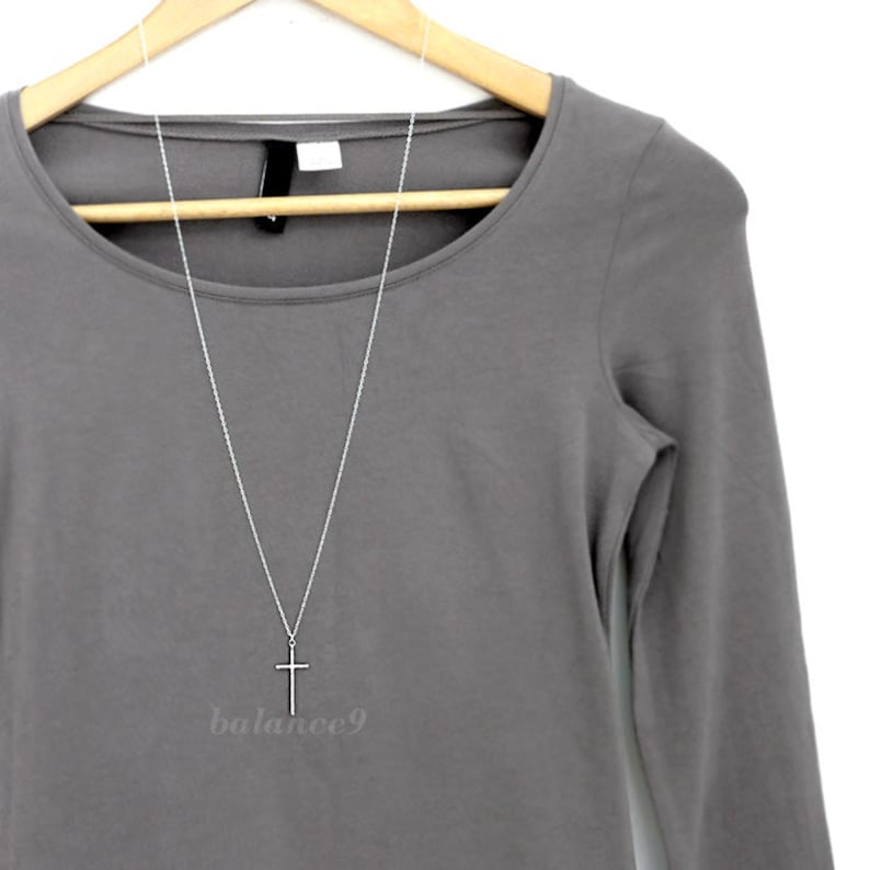 May include: A silver chain necklace with a simple silver cross pendant hanging from it. The necklace is displayed on a grey long-sleeved shirt.