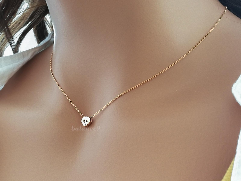 Puede incluir: Un delicado collar dorado con un peque&ntilde;o colgante de calavera. La calavera tiene detalles de ojos y nariz. El collar se muestra en el cuello de una persona, mostrando el tama&ntilde;o y el dise&ntilde;o del colgante. La cadena es fina y elegante.