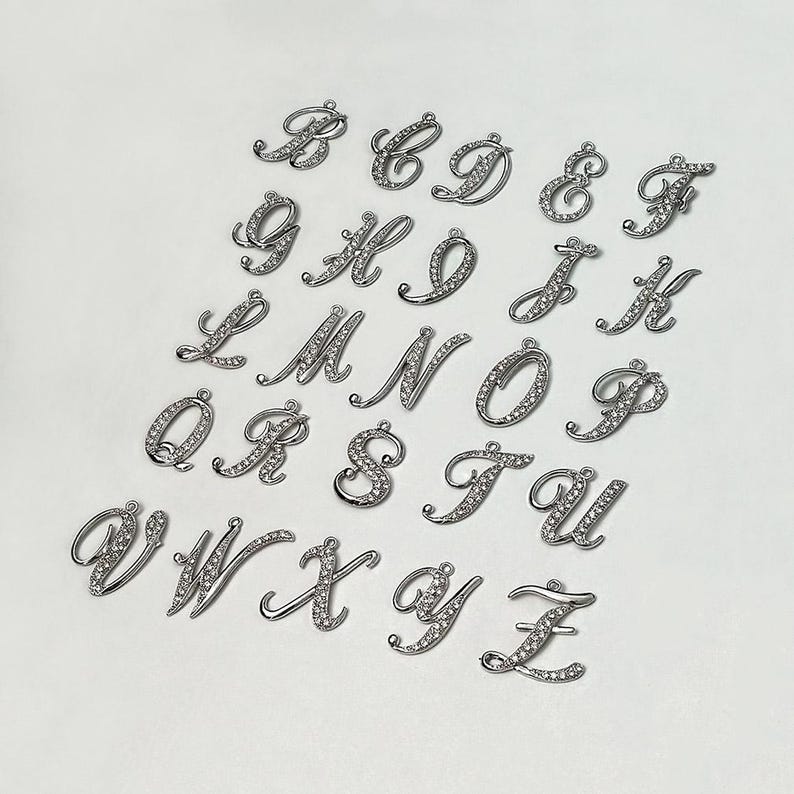 May include: A set of 26 silver-toned metal alphabet letter charms, each letter is encrusted with clear rhinestones. The letters are arranged in alphabetical order.