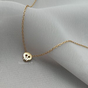Puede incluir: Un collar dorado con un peque&ntilde;o colgante de calavera. La calavera tiene dos cuencas de los ojos y una cavidad nasal. La cadena del collar es delicada y parece estar hecha de peque&ntilde;os eslabones conectados. El fondo es una tela gris claro y suave.