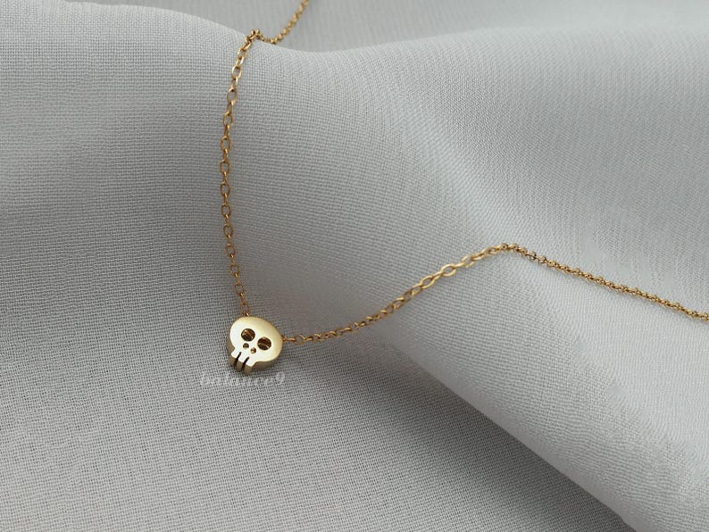 Puede incluir: Un collar dorado con un peque&ntilde;o colgante de calavera. La calavera tiene dos cuencas de los ojos y una cavidad nasal. La cadena del collar es delicada y parece estar hecha de peque&ntilde;os eslabones conectados. El fondo es una tela gris claro y suave.