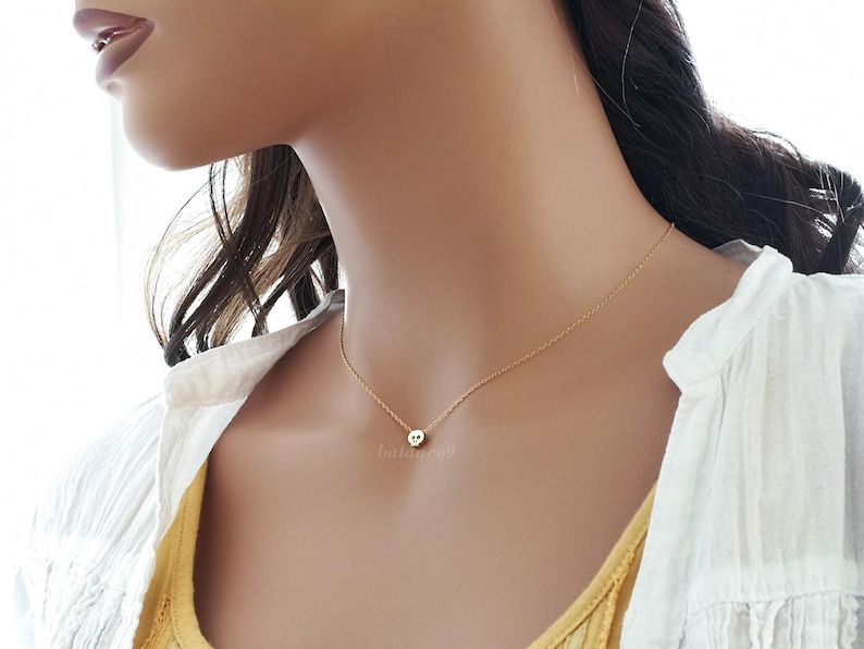 Puede incluir: Delicado collar dorado con un peque&ntilde;o colgante. El colgante tiene un dise&ntilde;o de calavera. El collar se lleva en el cuello de una persona, sobre una camiseta amarilla y una camisa blanca.