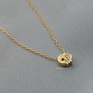 Puede incluir: Un collar dorado con un peque&ntilde;o colgante de calavera. La calavera tiene recortes para los ojos y la nariz y est&aacute; unida a una delicada cadena. La joya se exhibe sobre un fondo gris.