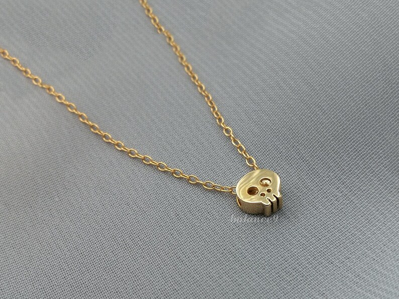 Puede incluir: Un collar dorado con un peque&ntilde;o colgante de calavera. La calavera tiene recortes para los ojos y la nariz y est&aacute; unida a una delicada cadena. La joya se exhibe sobre un fondo gris.