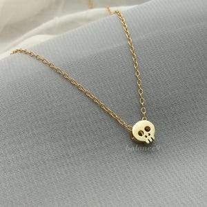 Puede incluir: Un delicado collar dorado con un peque&ntilde;o colgante de calavera. La calavera tiene dos cuencas de los ojos y una cavidad nasal. La cadena es fina y el collar se muestra sobre una superficie gris.