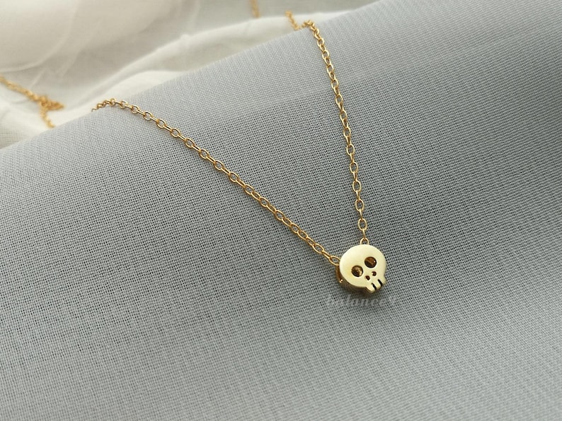 Puede incluir: Un delicado collar dorado con un peque&ntilde;o colgante de calavera. La calavera tiene dos cuencas de los ojos y una cavidad nasal. La cadena es fina y el collar se muestra sobre una superficie gris.
