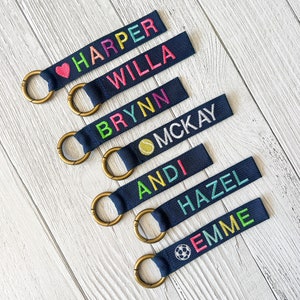 May include: Navy blue keychains with embroidered names in rainbow colors. Each keychain has a gold-colored ring. Some keychains feature a heart, tennis ball, and soccer ball icon next to the name.