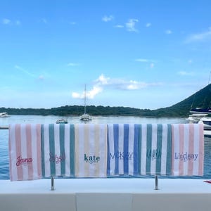 May include: Six striped beach towels hanging in a row, each featuring a unique embroidered name and the text BVI 2024 with an anchor icon. The towels are coloured in soft pink, teal, yellow, and blue stripes, displayed against a bright blue sky.