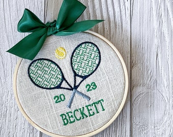Puede incluir: Un adorno de tenis bordado en blanco y verde con el nombre "BECKETT" y los números "20" y "23" sobre un fondo de lino natural. El adorno está sujeto a un lazo de satén verde.