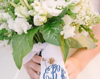 May include: A white bridal bouquet with green leaves and a white ribbon tied around the stems. The ribbon has a blue monogram and the date "4.5.14" printed on it.