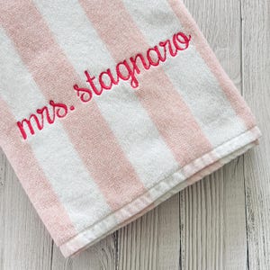 May include: A soft, plush towel featuring vertical stripes in alternating shades of light pink and white. The fabric is embroidered with the text "mrs. stagnaro" in a vibrant, dark pink cursive script, positioned diagonally across the striped pattern.