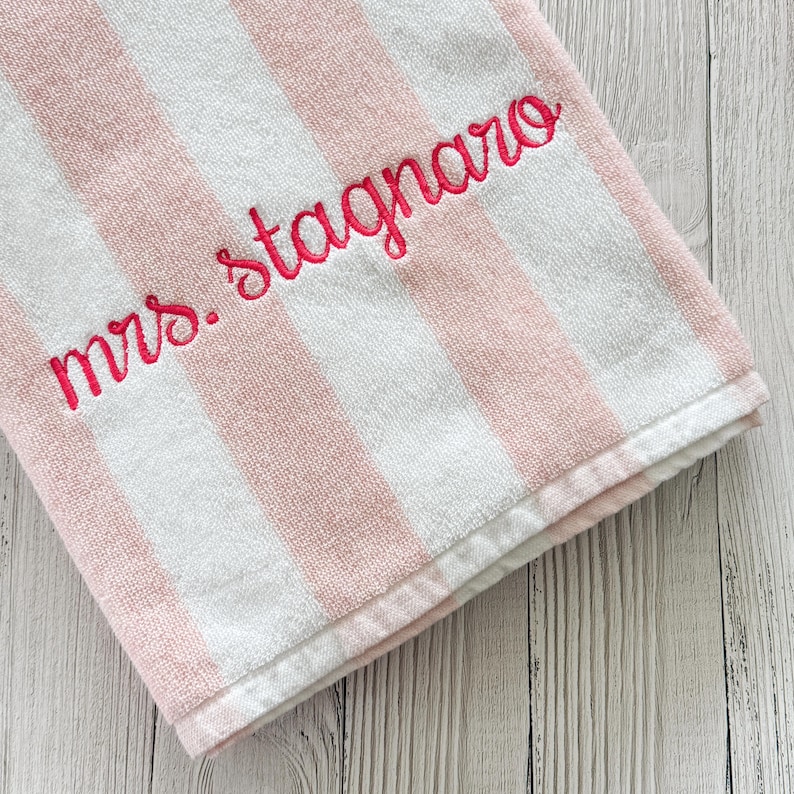 May include: A soft, plush towel featuring vertical stripes in alternating shades of light pink and white. The fabric is embroidered with the text "mrs. stagnaro" in a vibrant, dark pink cursive script, positioned diagonally across the striped pattern.