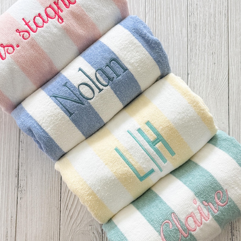 May include: Four rolled beach towels featuring vertical stripes in pastel pink, blue, yellow, and green, each paired with white. The towels are personalised with embroidered names and initials in matching thread colours, including s.stagn, Nolan, LH, and Claire.