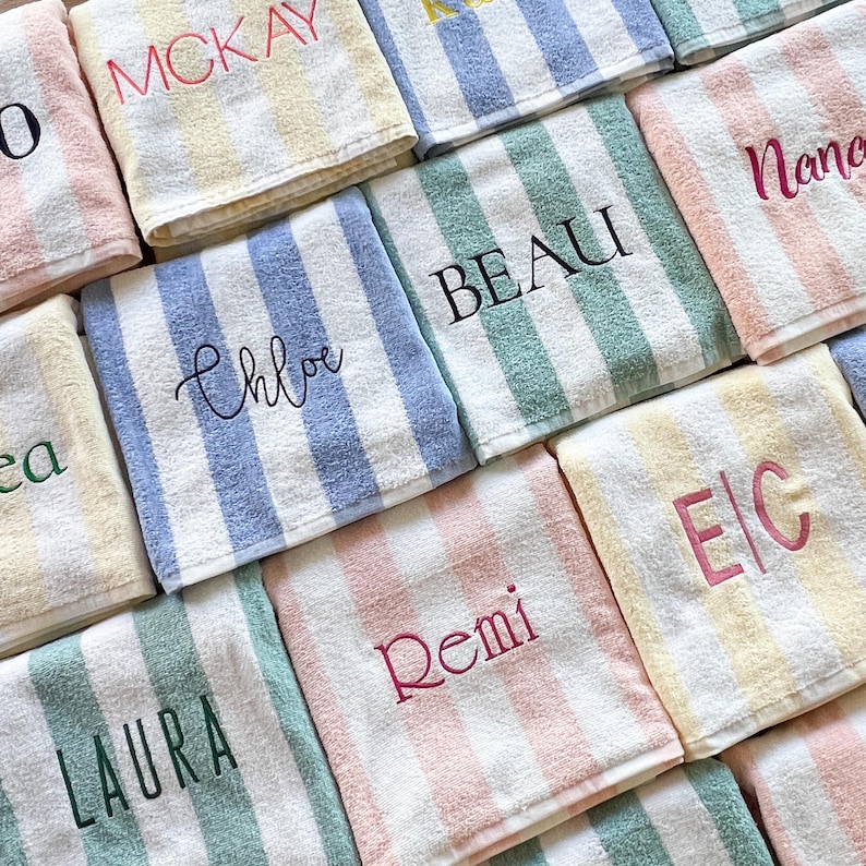May include: A collection of folded rectangular towels featuring vertical stripes in pastel colours including yellow, blue, green, and pink. Each towel is embroidered with a unique name or initials in various fonts and thread colours.