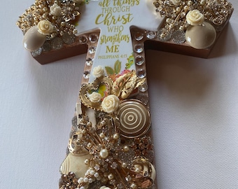 May include: A white wooden cross decorated with gold and white embellishments, including buttons, beads, and flowers. The cross features the text "I can do all things through Christ who strengthens me. Philippians 4:13."