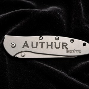 May include: A silver folding knife with the name "AUTHUR" engraved on the handle. The brand name "kershaw" is also visible. The knife is set against a dark, textured background.