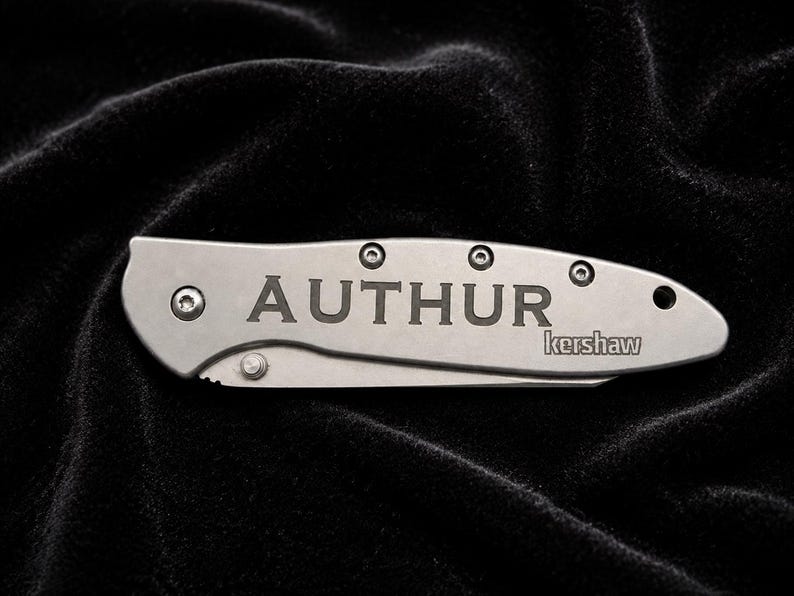 May include: A silver folding knife with the name "AUTHUR" engraved on the handle. The brand name "kershaw" is also visible. The knife is set against a dark, textured background.