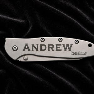 May include: A silver pocket knife with the name "ANDREW" engraved on the blade. The brand name "kershaw" is also visible. The knife is set against a black, textured background.
