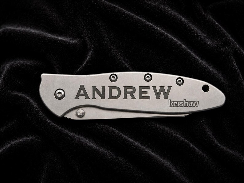May include: A silver pocket knife with the name "ANDREW" engraved on the blade. The brand name "kershaw" is also visible. The knife is set against a black, textured background.