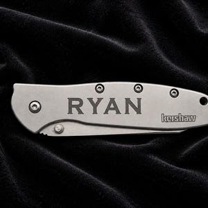 May include: A silver folding knife with the name "RYAN" engraved on the handle. The brand name "kershaw" is also visible. The knife is set against a black, textured background.