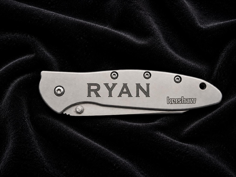 May include: A silver folding knife with the name "RYAN" engraved on the handle. The brand name "kershaw" is also visible. The knife is set against a black, textured background.