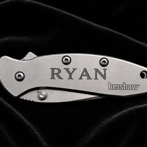 May include: A silver folding knife with the name "RYAN" engraved on the handle. The brand name "kershaw" is also visible. The knife is on a black background.