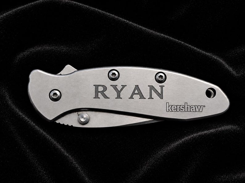 May include: A silver folding knife with the name "RYAN" engraved on the handle. The brand name "kershaw" is also visible. The knife is on a black background.
