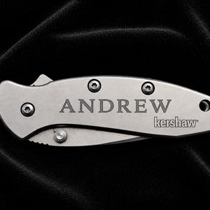 May include: A silver folding knife with the name "ANDREW" engraved on the handle. The brand name "kershaw" is also visible. The knife is shown against a black background.