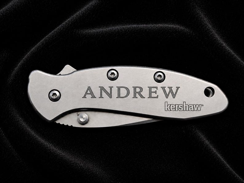 May include: A silver folding knife with the name "ANDREW" engraved on the handle. The brand name "kershaw" is also visible. The knife is shown against a black background.