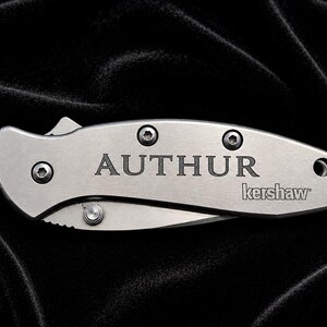 May include: A silver folding knife with the word "AUTHOR" engraved on the handle. The brand name "kershaw" is also visible. The knife is set against a black background.