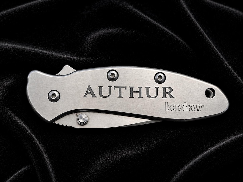 May include: A silver folding knife with the word "AUTHOR" engraved on the handle. The brand name "kershaw" is also visible. The knife is set against a black background.