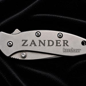 May include: A silver folding knife with the name "ZANDER" engraved on the handle, along with the "kershaw" brand. The knife has a curved blade and is set against a dark, textured background.