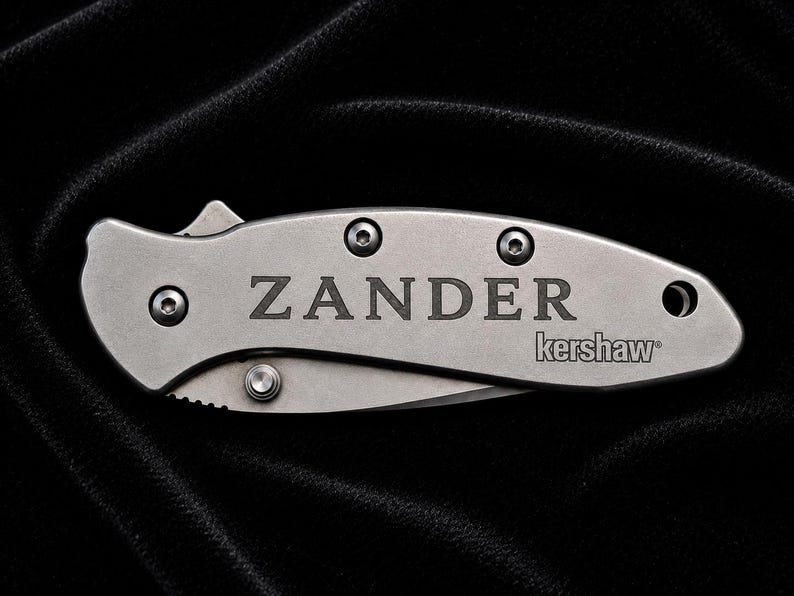 May include: A silver folding knife with the name "ZANDER" engraved on the handle, along with the "kershaw" brand. The knife has a curved blade and is set against a dark, textured background.