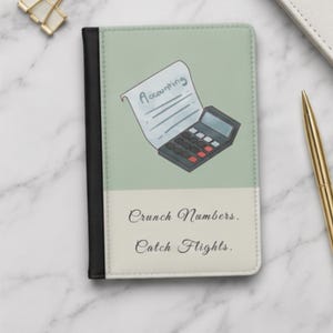 Puede incluir: Una funda para pasaporte con lomo negro y cubierta de dos tonos. La parte superior es verde salvia con una ilustración de una calculadora y un papel que dice Accounting. La parte inferior es crema con el texto Crunch Numbers. Catch Flights.