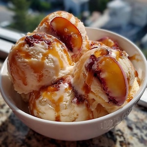 May include: A white bowl of vanilla ice cream scoops, drizzled with caramel sauce and peach slices. The ice cream is creamy, the peaches are orange and red. The bowl is on a surface with a blurred background. The image is well-lit.