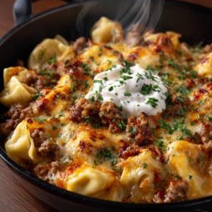 Könnte beinhalten: Nahaufnahme eines gebackenen Tortellini-Gerichts in einer schwarzen Gusseisenpfanne. Das Gericht besteht aus mit Käse bedeckten Tortellini, Hackfleisch und einem Klecks weißer Sahne, garniert mit gehackter Petersilie. Dampf steigt von dem heißen Essen auf.