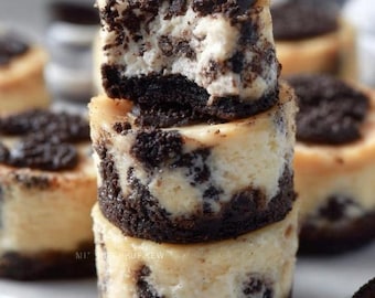 May include: A stack of three mini cheesecakes with Oreo cookie crusts and toppings. The creamy, off-white filling is speckled with dark cookie pieces. One cheesecake has a bite taken out of it, revealing the interior.