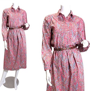 May include: A long-sleeved, collared dress with a paisley pattern in shades of red, blue, and brown. The dress features a pleated bodice, a brown belt, and a midi-length skirt. The dress is displayed on a mannequin.