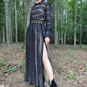 May include: A black lace maxi dress with long sleeves, a high neckline, and a thigh-high slit. The dress has a fitted bodice and a flowing skirt. The model is wearing the dress with black ankle boots.