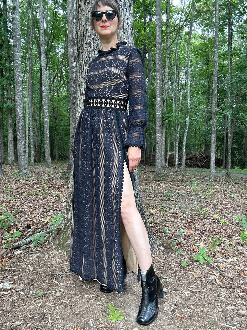 May include: A black lace maxi dress with long sleeves, a high neckline, and a thigh-high slit. The dress has a fitted bodice and a flowing skirt. The model is wearing the dress with black ankle boots.