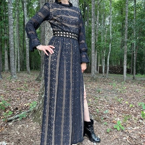 May include: A black lace maxi dress with a high neck, long sleeves, and a fitted waist. The dress has a black belt with a geometric design and a thigh-high slit. The model is wearing black boots.